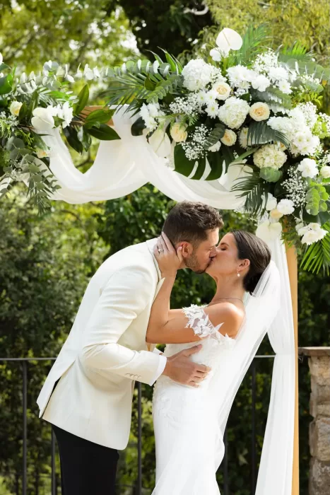 Couple sharing their first kiss under a white floral arch at Mas del Sord.