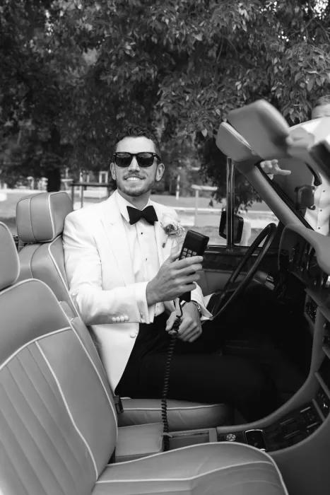 Groom sitting in the vintage convertible at Mas del Sord, holding the retro car phone with a smile.