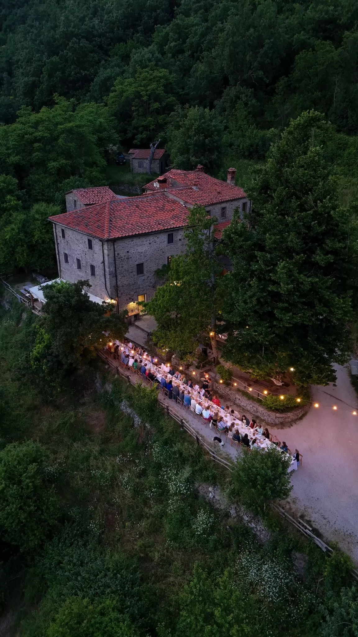 Aerial view of long dinner table lit by string lights outside the stone farmhouse at Novanta 90