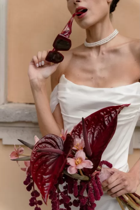 Chic bride holding bold red bouquet and red sunglasses with a pearl necklace at Villa Buttafava.