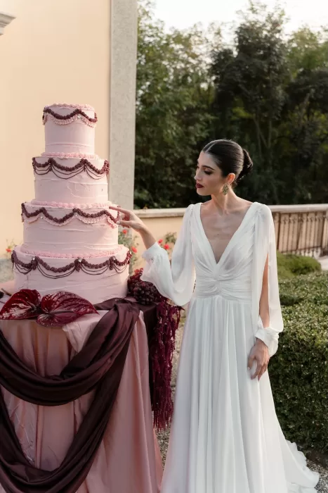 Bride in elegant white gown admiring a pink tiered wedding cake with burgundy accents at Villa Buttafava.