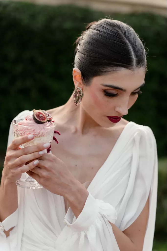 Bride in white gown holding a pink fig dessert in an elegant coupe glass at Villa Buttafava.