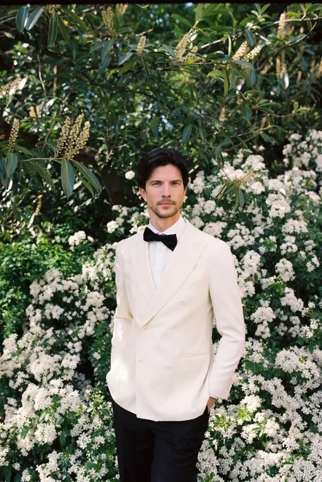 Groom in an ivory tuxedo standing before lush white blooms in the gardens of Château du Prieuré.