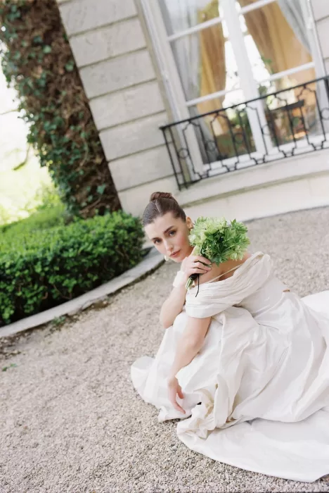 Bride seated on gravel with a green bouquet, leaning softly against her shoulder outside Château du Prieuré.