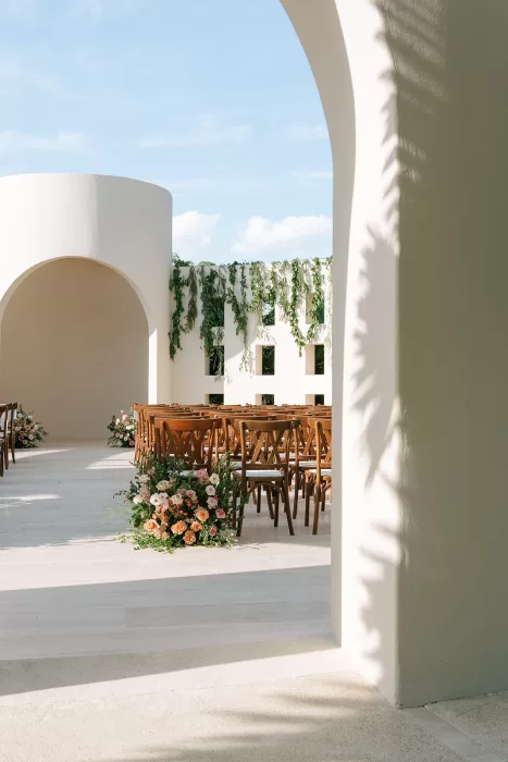 Wedding ceremony aisle at Baja Luna viewed through an arched walkway, with wooden chairs and floral arrangements.