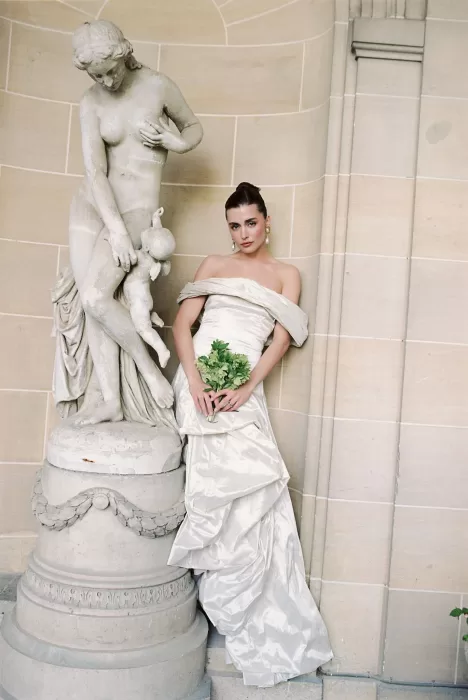 Bride posing beside a classical stone statue in the arched exterior of Château du Prieuré, holding a simple green bouquet.
