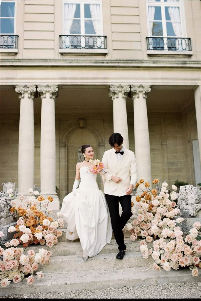 Bride and groom walking down the floral-lined steps of Château du Prieuré, surrounded by lush pastel roses and grand stone columns.