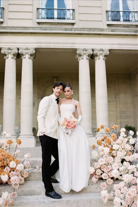 Bride and groom standing together on the floral-lined steps of Château du Prieuré, framed by grand stone columns.