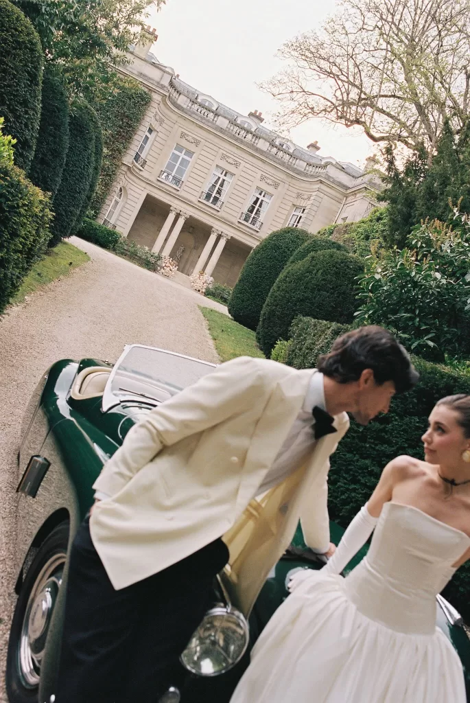 Couple sharing a playful moment beside a vintage car on the tree-lined driveway of Château du Prieuré.