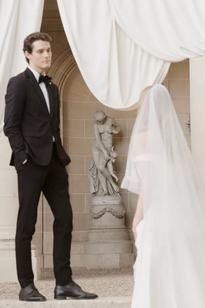 Bride and groom meeting beneath soft drapery near a stone statue at Château du Prieuré d’Evecquemont.
