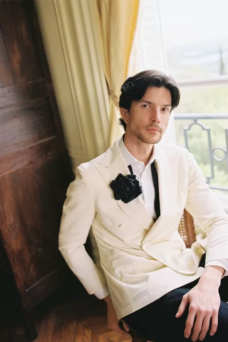 Groom seated by a tall window at Château du Prieuré, wearing an ivory tuxedo with a black floral lapel accent.