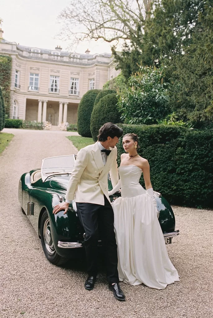 Stylish couple leaning against a vintage green car in front of Château du Prieuré, capturing an elegant and romantic arrival moment.