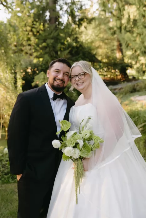 Bride and groom smile together in the gardens at Chateau Lill, holding a white and green bouquet in warm afternoon light.