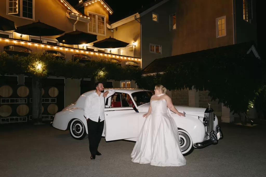 Bride and groom pose beside a vintage white car at night, illuminated by warm lights in the courtyard of Chateau Lill.