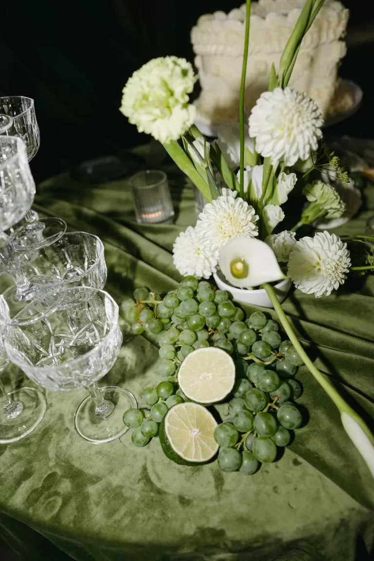 Styled wedding still life with champagne coupes, green grapes, citrus and white florals on velvet linens at Chateau Lill.