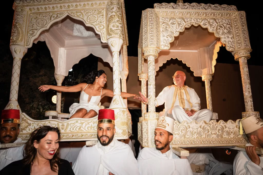 Bride and groom laughing as they are carried in ornate traditional hammam litters during a lively celebration at Jnane Tamsna.