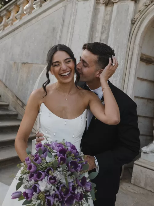 Joyful portrait of the bride and groom holding a purple bouquet on the stone steps at Vandelli Botanical Garden.