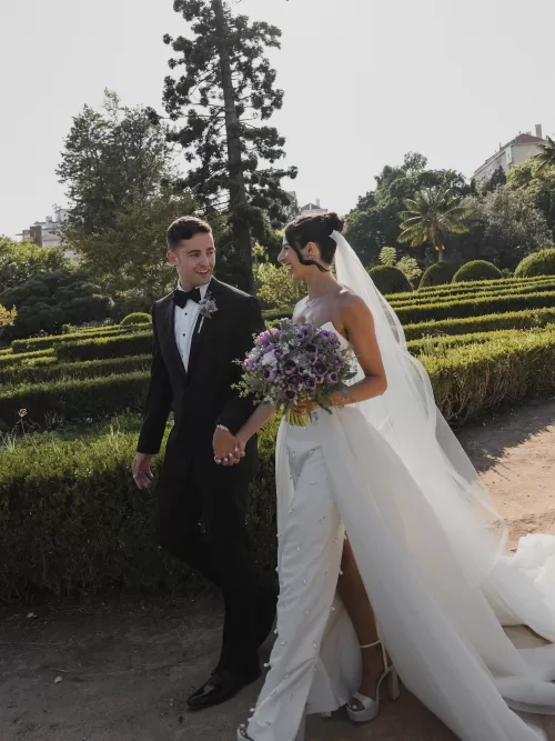 Newlyweds walking hand in hand through manicured hedges with a purple bouquet at Vandelli Botanical Garden.