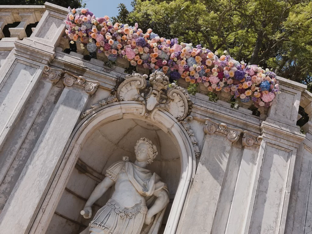 Bride and groom posing beside classical stone architecture adorned with floral installations at Vandelli Botanical Garden.