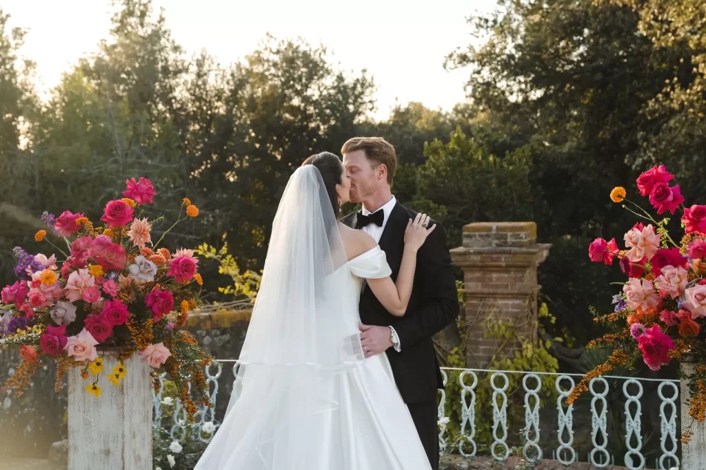 The couple shares a kiss at the Villa Lena altar, framed by vibrant pink and orange floral arrangements.