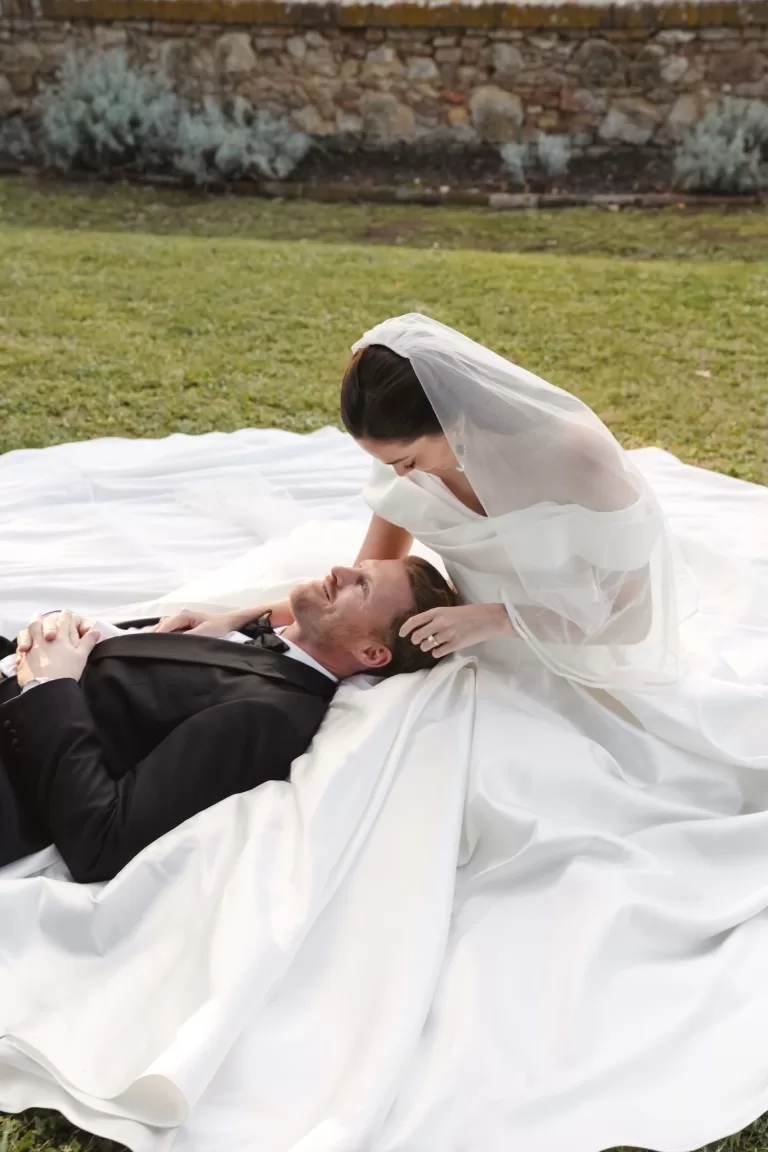 The bride kneels beside her partner as he lies on her gown’s sweeping train during portraits at Villa Lena.