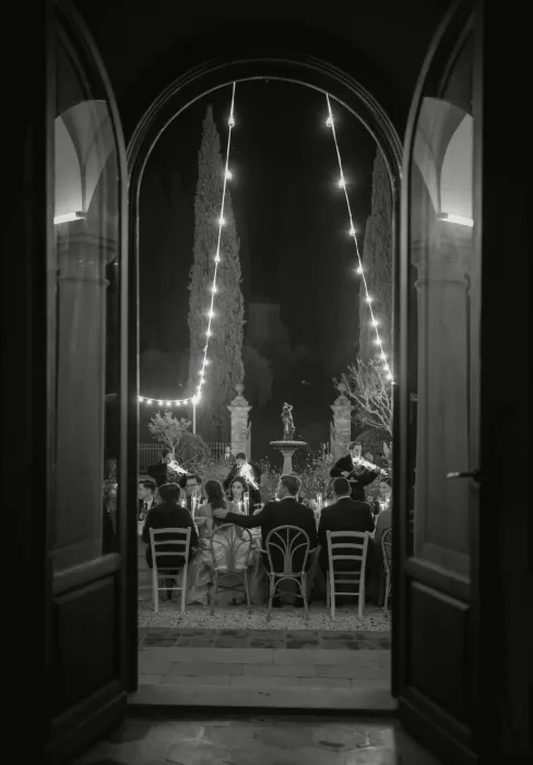 Nighttime view through arched doors framing the Villa Lena courtyard dinner, lit with string lights and live musicians.