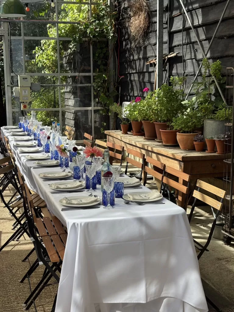 Long white-linen reception table inside the Worton Kitchen Garden glasshouse, styled with blue glassware and potted herbs.