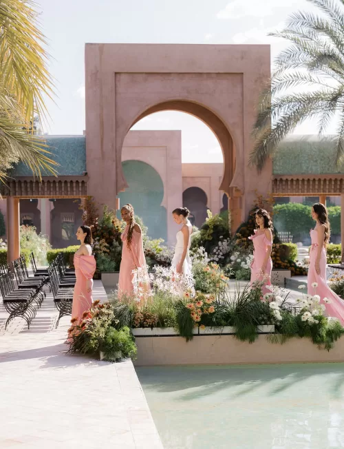 Bridesmaids in soft pink dresses walking along the poolside ceremony walkway at an Amanjena wedding.