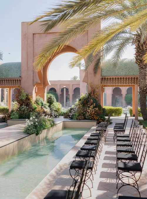 Poolside wedding ceremony at Amanjena with black chairs lining the water and lush floral arrangements beneath palm trees.