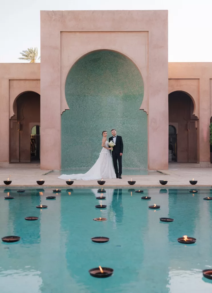 Newlyweds standing beside the reflecting pool at Amanjena, framed by the iconic tiled arch and floating candlelight.