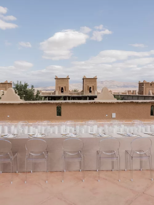 Long wedding dinner table on the Amanjena terrace with clear chairs, neutral linens and views across the Atlas foothills.