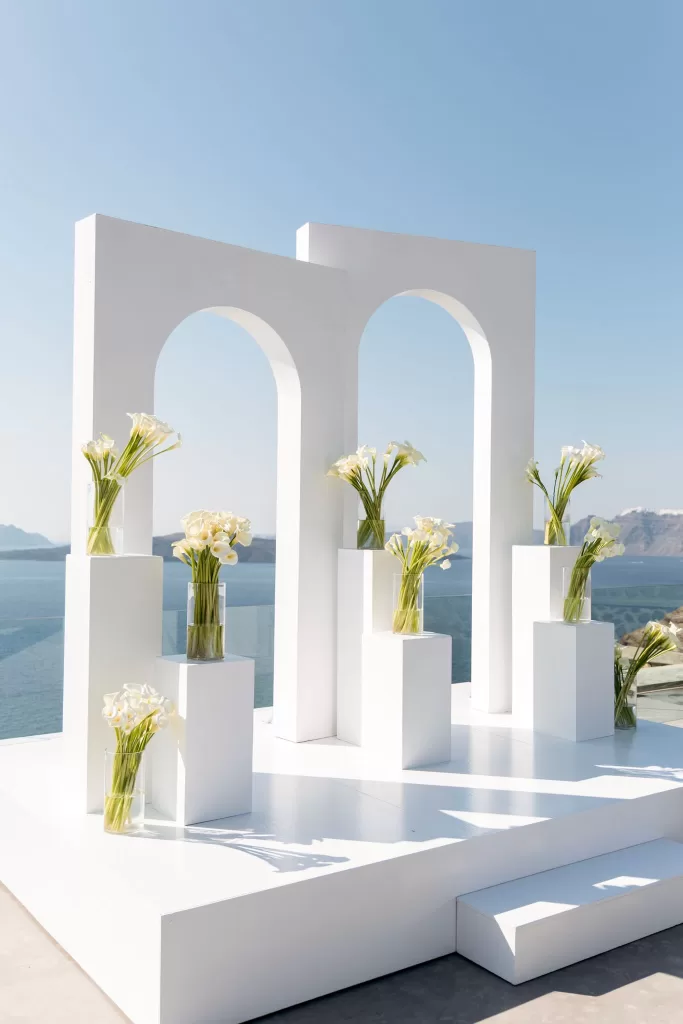 Minimalist white wedding ceremony arches at Cavo Ventus styled with calla lilies, set against panoramic views of the Aegean Sea.