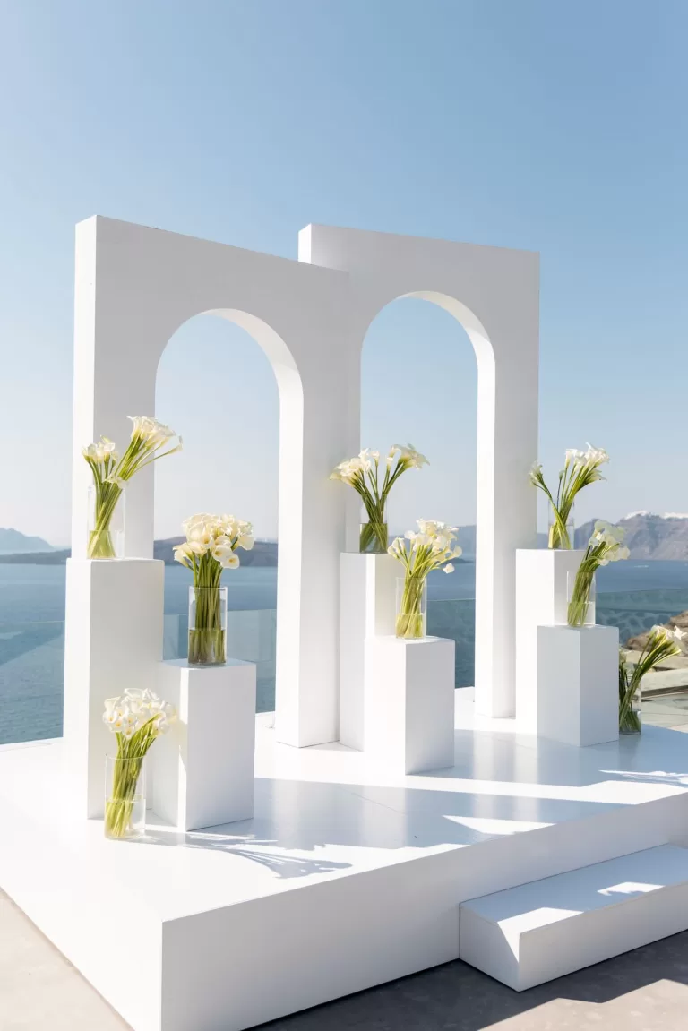 Minimalist white wedding ceremony arches at Cavo Ventus styled with calla lilies, set against panoramic views of the Aegean Sea.