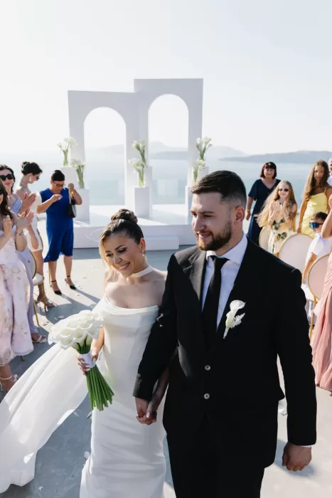 Newlyweds walking hand in hand down the aisle at Cavo Ventus, surrounded by guests and minimalist white arches overlooking the Aegean Sea.
