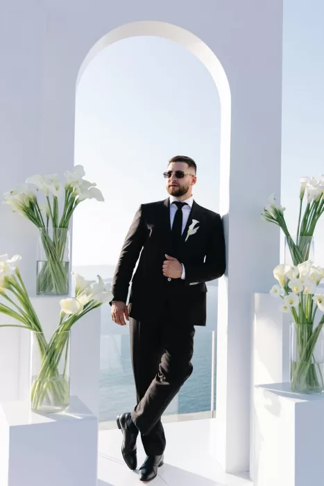 Groom standing beneath a white ceremony arch at Cavo Ventus, styled with calla lilies and overlooking the Aegean Sea.