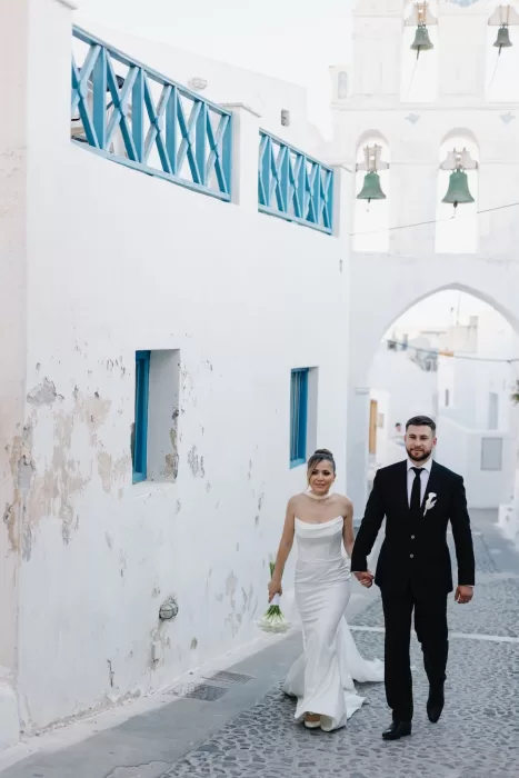 Bride and groom walking through a traditional whitewashed Santorini street near Cavo Ventus, framed by blue details and church bells.