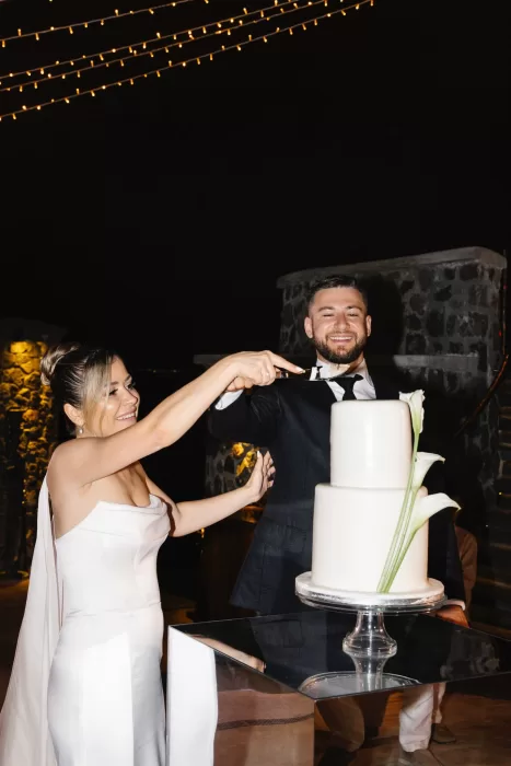 Bride and groom cutting a modern white wedding cake at Cavo Ventus under warm evening lights, with volcanic stone walls creating an intimate night-time atmosphere.