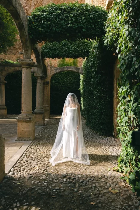 Bride standing beneath stone arches in the cloister walkway at Finca Es Convent, surrounded by greenery.