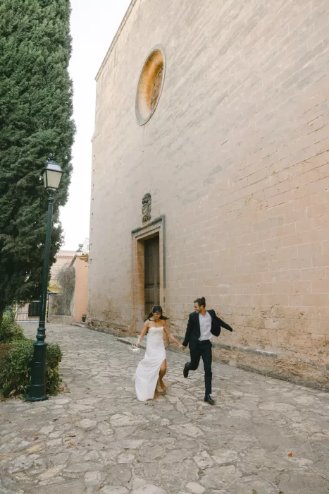 Newlyweds running hand in hand along a quiet stone street beside Finca Es Convent after their wedding.