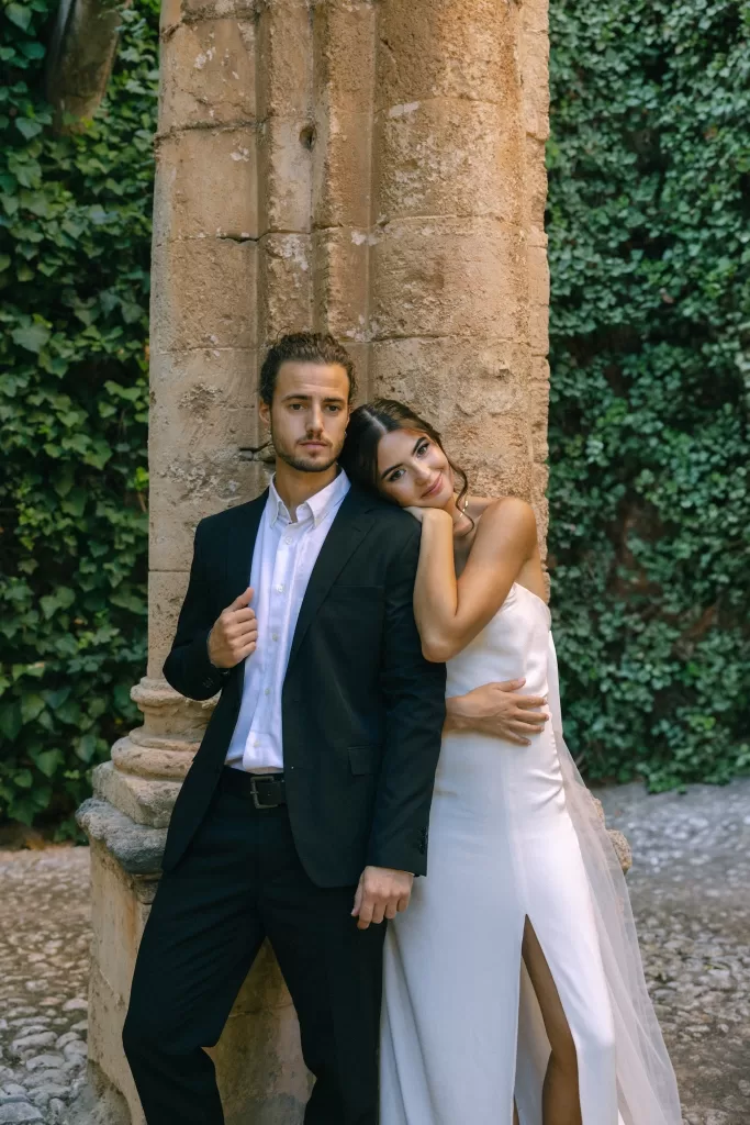 Bride leaning into her groom beside a historic stone column at Finca Es Convent, framed by ivy-covered walls.