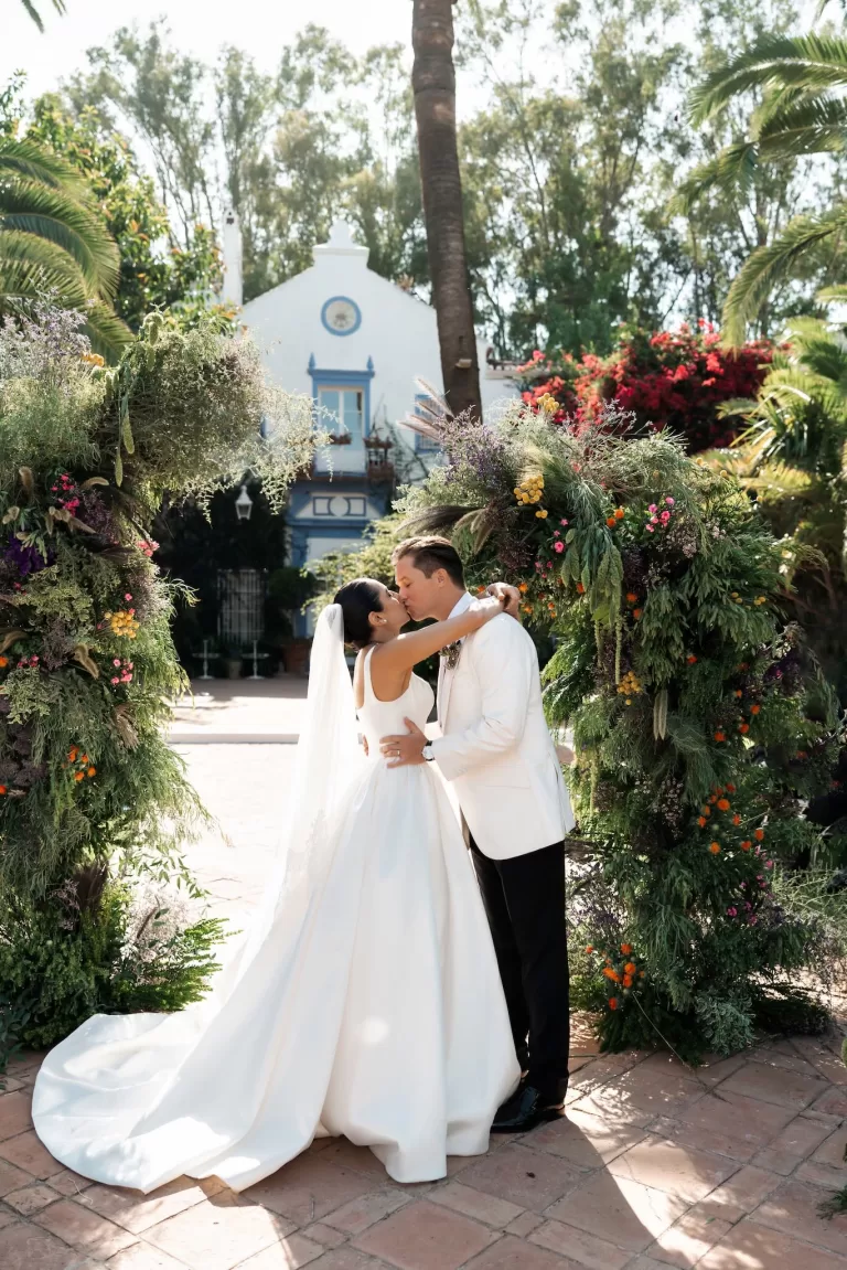Bride and groom kissing beneath a lush floral ceremony arch in the courtyard of Finca La Concepción.