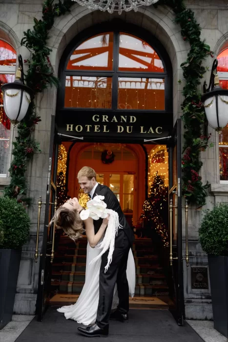Bride and groom kissing beneath the entrance of Grand Hotel du Lac, framed by festive lights and winter greenery.