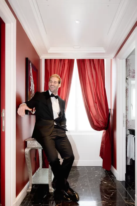 Groom in a black tuxedo relaxed in a red-toned suite at Grand Hotel du Lac, capturing a confident and playful pre-ceremony moment.