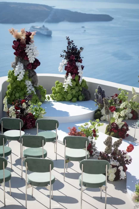 Modern wedding ceremony setup at Islet Santorini featuring sculptural floral installations and caldera views, with green velvet seating arranged around the altar.