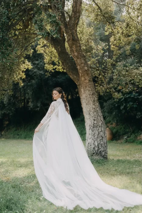 Bride wearing a flowing cape beneath the trees at Mas de Les Voltes, captured in a quiet and natural portrait.