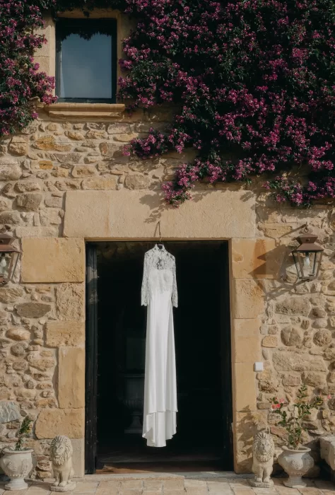 Elegant lace wedding dress displayed against the rustic stone walls of Mas de Les Voltes, framed by climbing flowers.