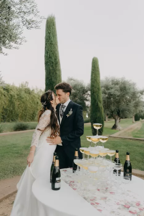Newlyweds sharing a kiss beside a champagne tower in the gardens of Mas de Les Voltes, framed by olive trees and cypress rows.