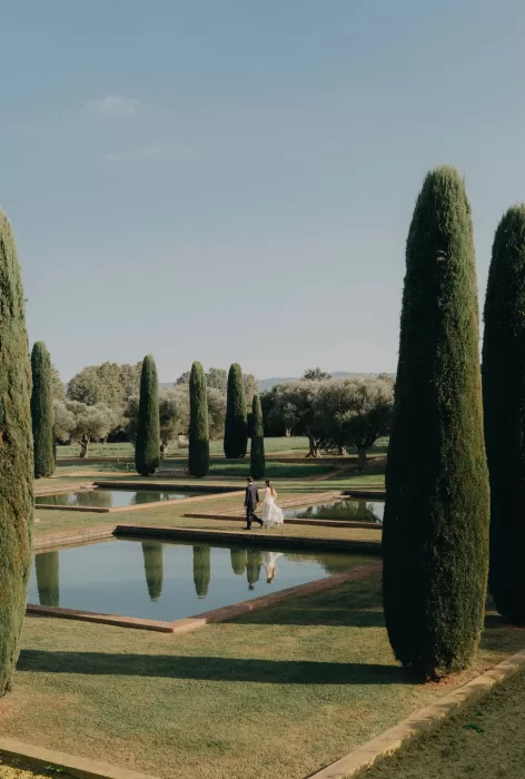 Newlyweds walking beside the reflecting pools and cypress trees in the gardens of Mas de Les Voltes under clear Mediterranean skies.