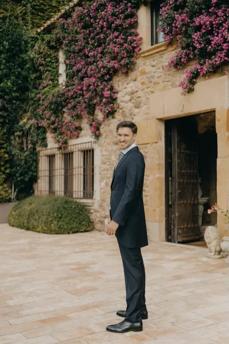 Groom portrait in the courtyard of Mas de Les Voltes, set against stone walls and flowering vines in soft natural light.