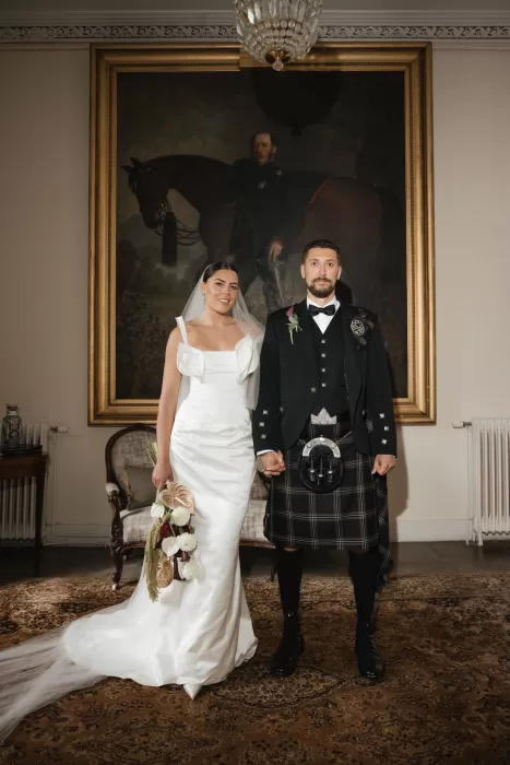 Bride and groom standing together beneath a large framed painting inside Boturich Castle.